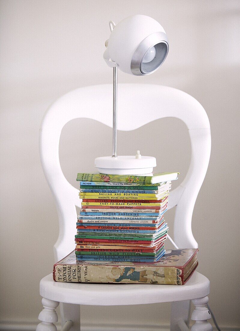 Stack of old childrens books with a contemporary lamp in white painted chair in Staffordshire home, England, UK