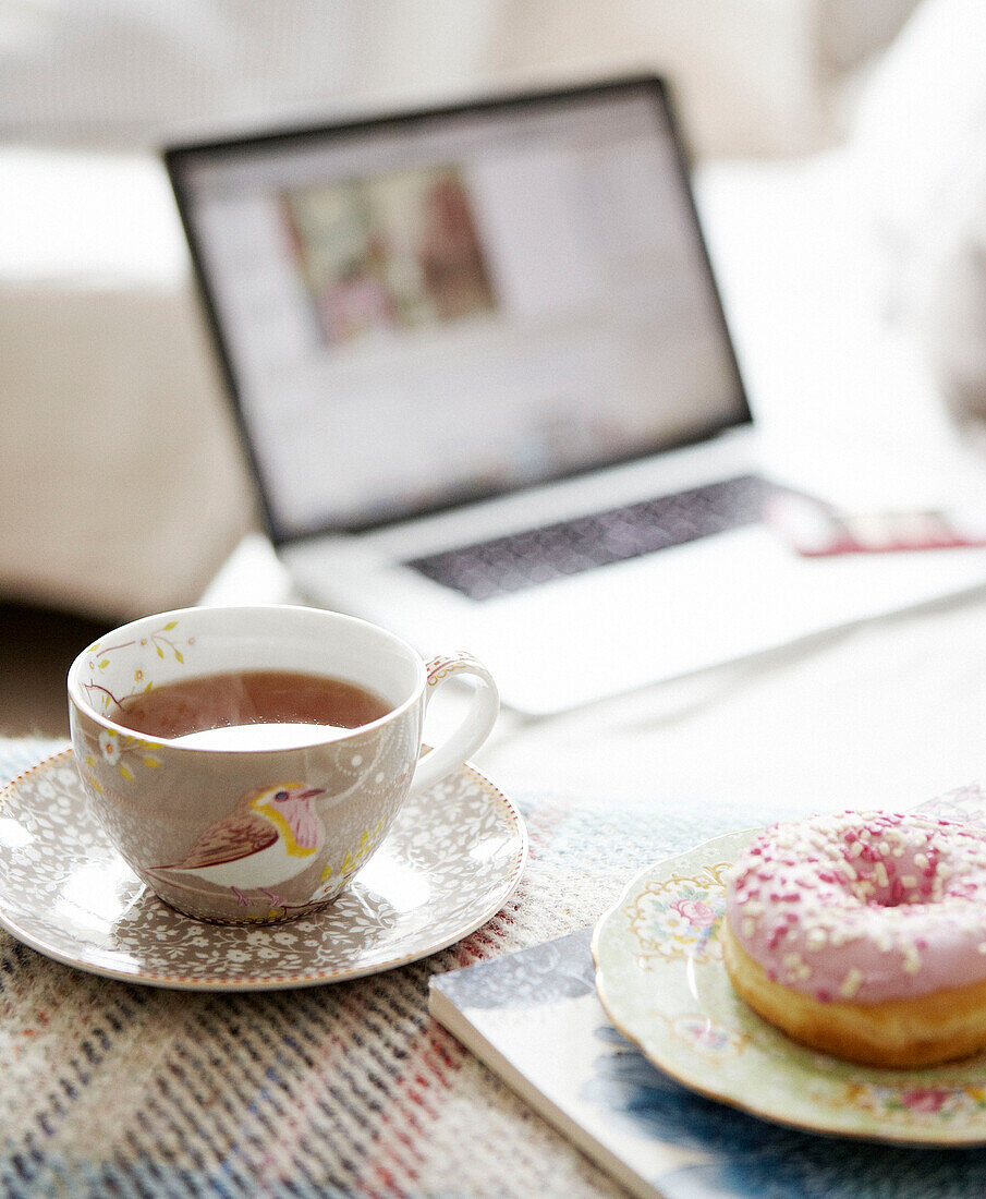 Hot tea and an iced ring donut with laptop computer in background in Hastings home, East Sussex, UK