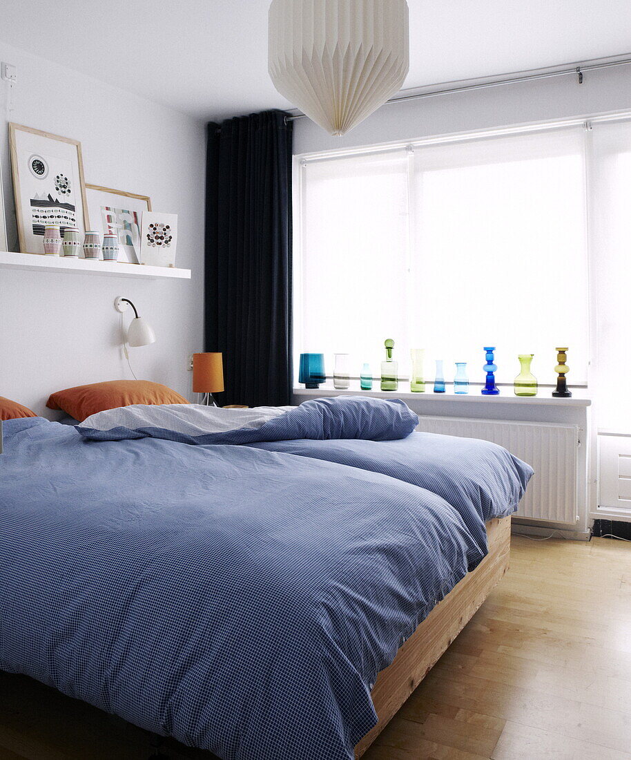 Retro glassware on windowsill of bedroom with blue checked duvet and shelf with artwork in Bussum home, Netherlands