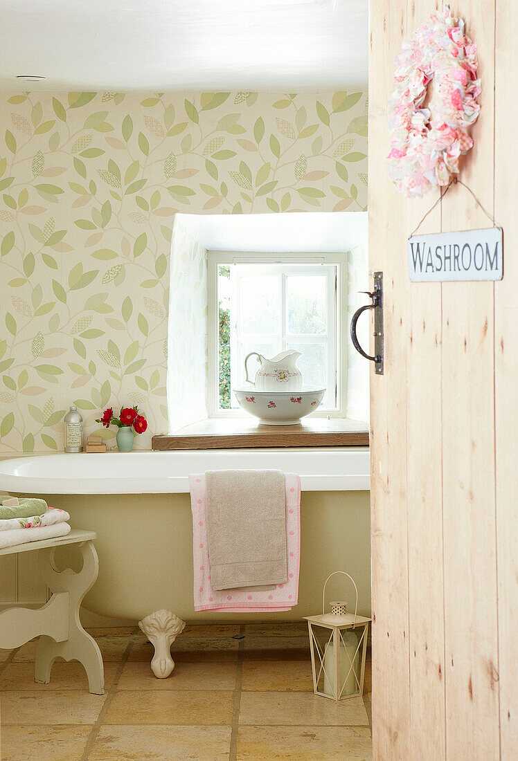 Freestanding bath below window with leaf motif wallpaper in Devonshire cottage UK
