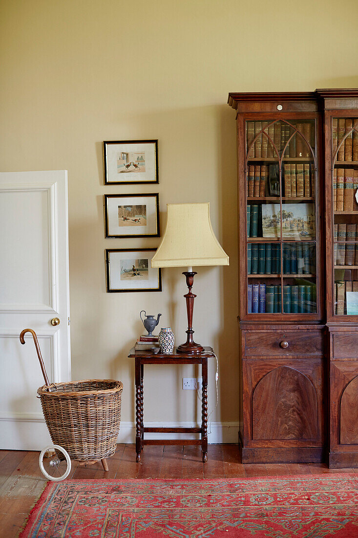 Yellow lamp on polished wooden side table with bookcase and shopping trolley in Capheaton Hall, Northumberland, UK