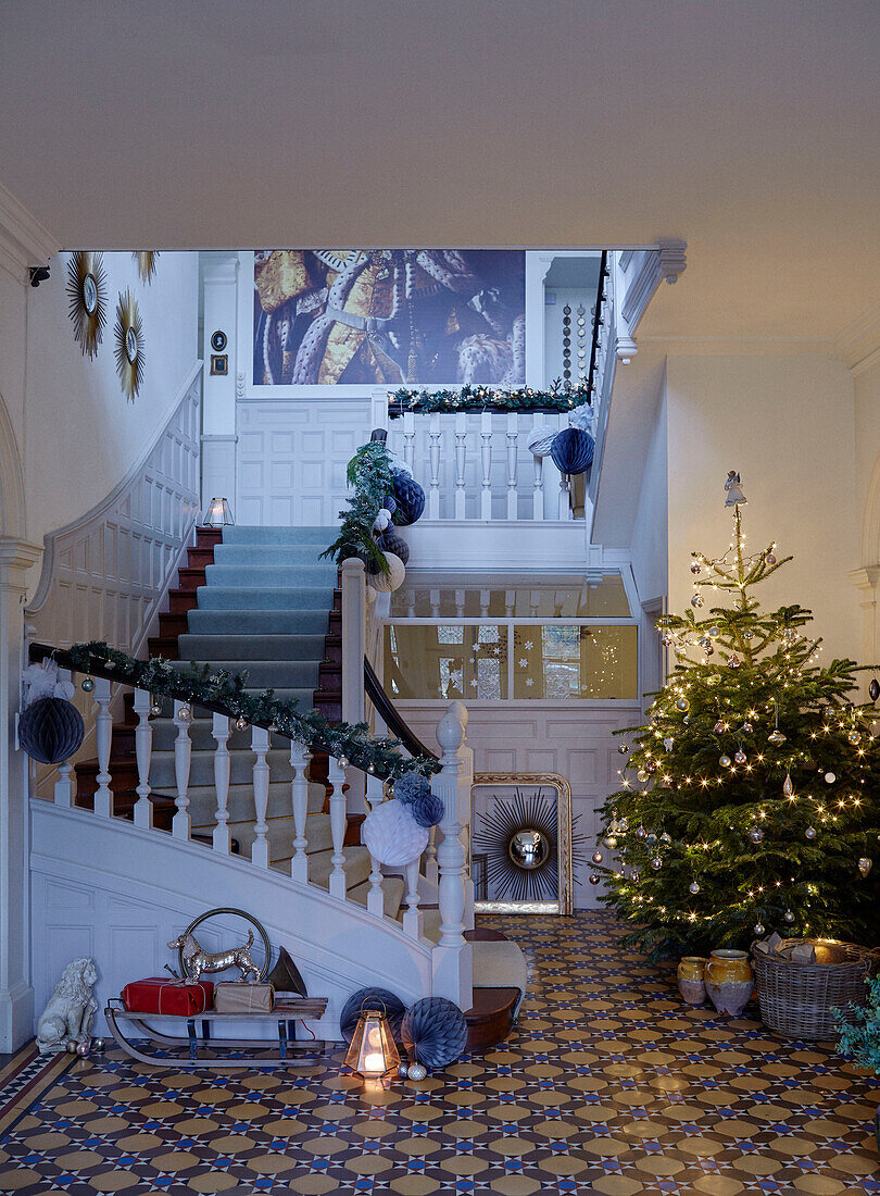 Girlanden am Geländer mit Weihnachtsbaum und Schlitten in der geräumigen gefliesten Eingangshalle eines Hauses in East Grinstead, West Sussex, UK