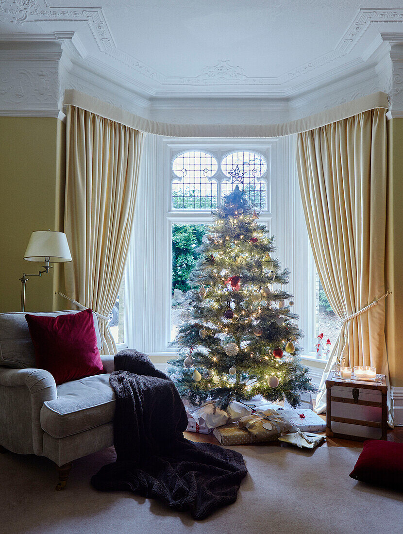 Weihnachtsgeschenke unter dem Baum im Erkerfenster mit Sessel und Decke in einem britischen Haus
