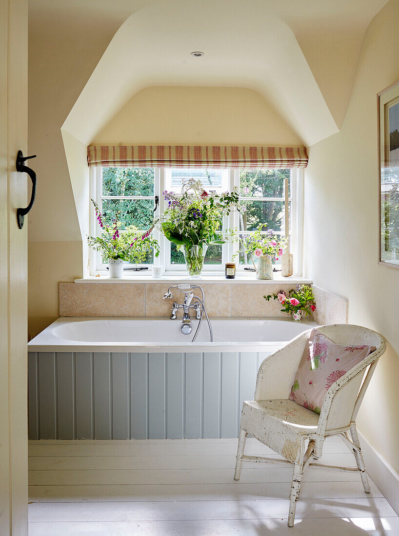 Cushion on chair with light blue panelled bath at window in Sandford St Martin cottage, Oxfordshire, UK