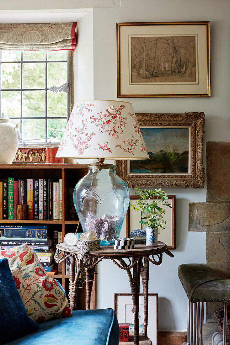 Glass lamp on side table with framed pictures and bookcase in Oxfordshire farmhouse, UK