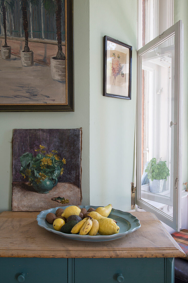 Fruit bowl and picture of flowers on top of old cabinet next to window