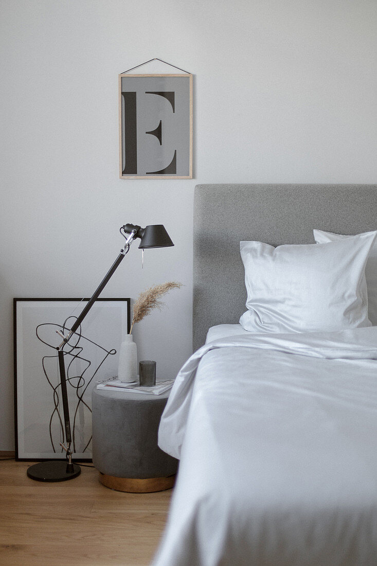 Bedside table and black lamp on floor next to bed with grey headboard and white bed linen