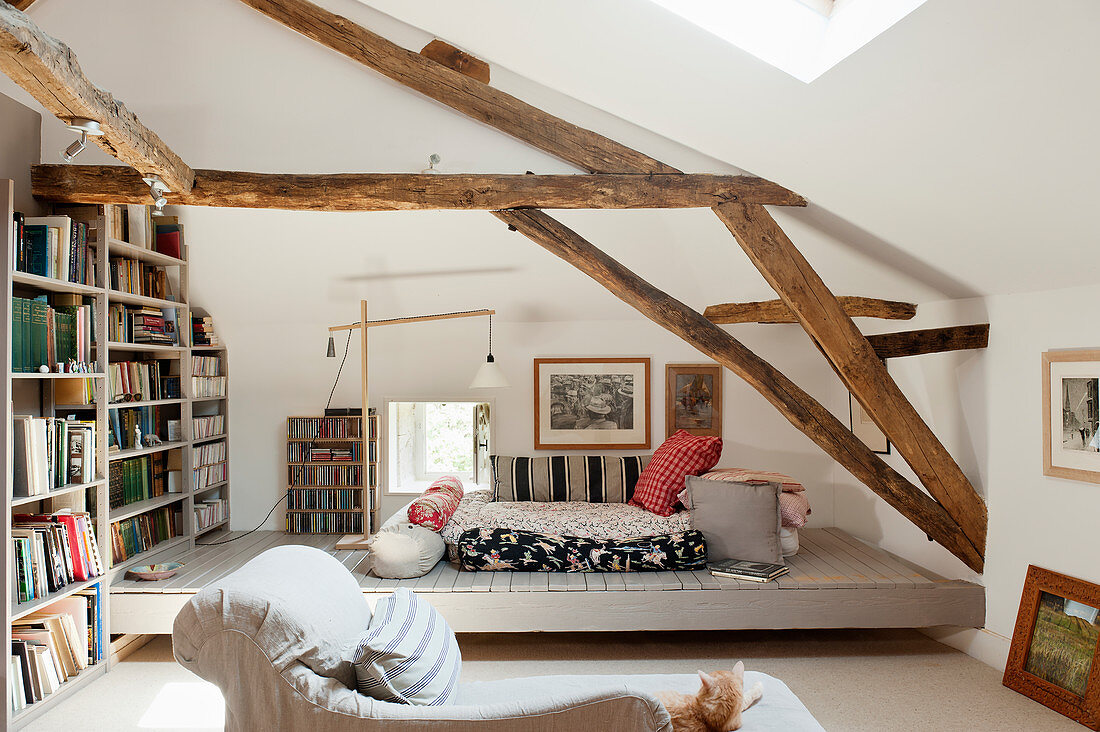 Dachzimmer mit Holzbalkendecken, großem Bücherregal, Bodenkissen und Chaiselongue