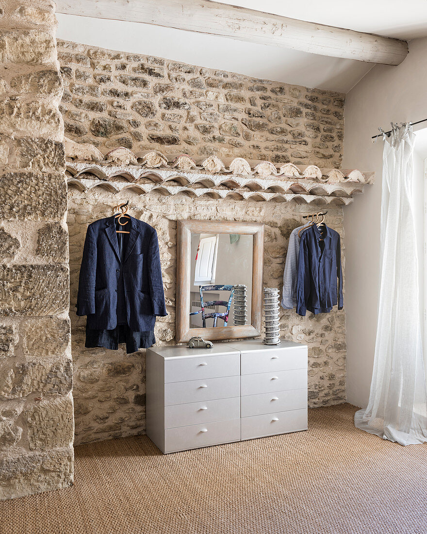 Clothes rails, chest of drawers and mirror in bedroom with stone wall