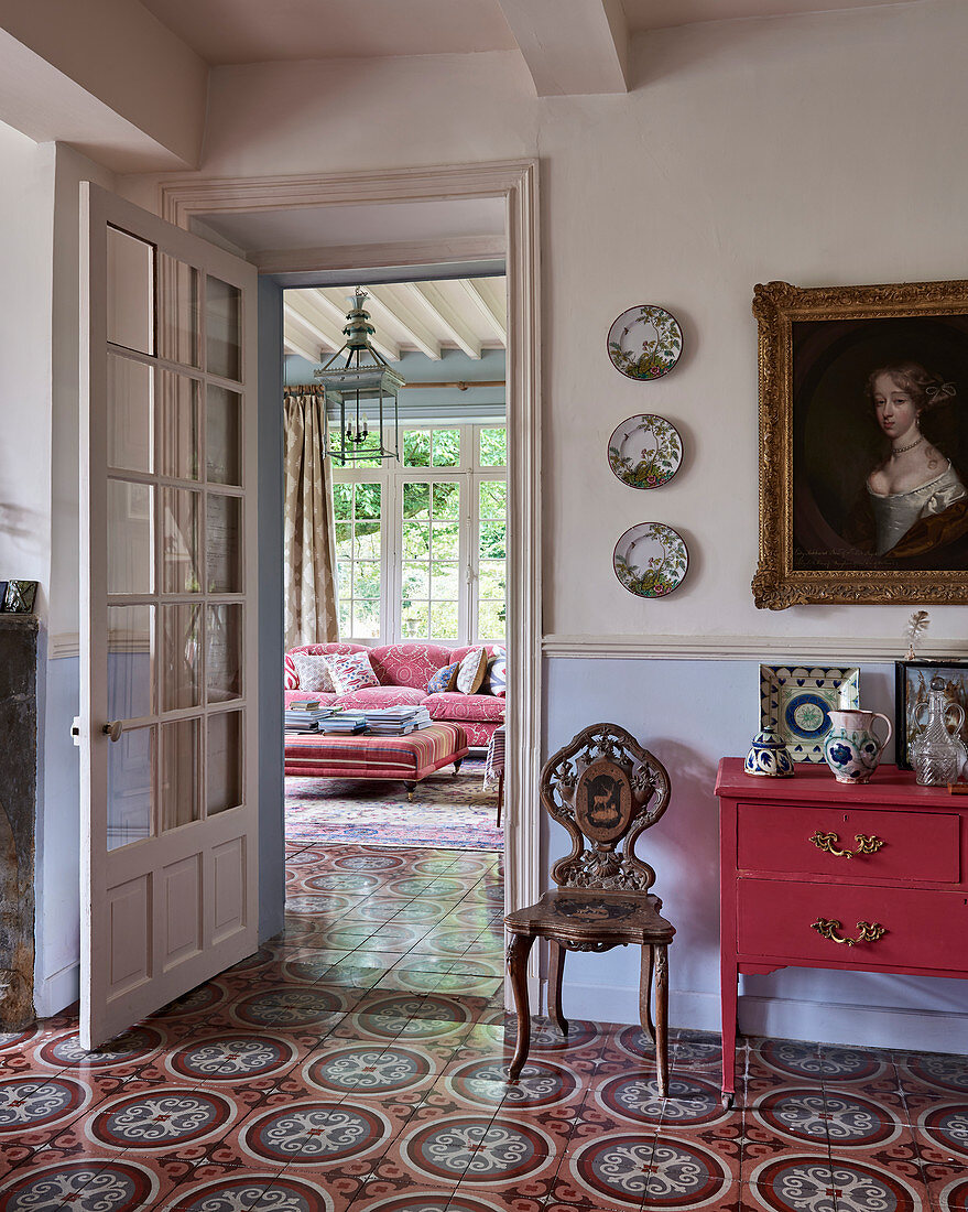 Red-painted cabinet and carved chair under portrait in hallway with decorative tiled floor