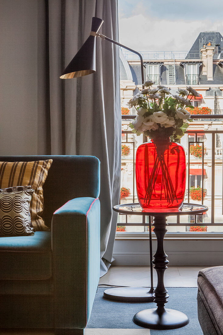 Rote Glasvase auf Beistelltisch und Stehlampe vor französischem Fenster
