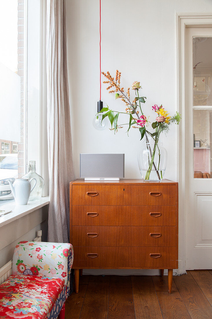 Flowers on retro chest of drawers below pendant lamp next to window