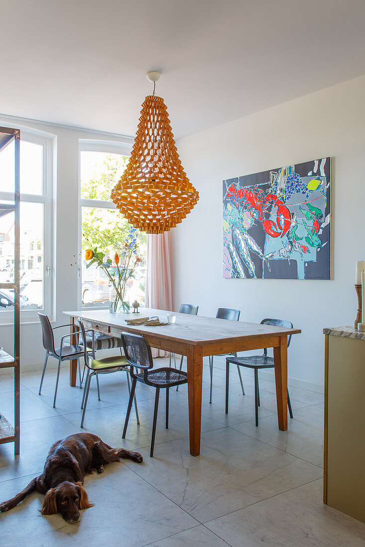 Pendant lamp above dining table and chairs next to modern artwork on wall; dog lying on tiled floor in foreground