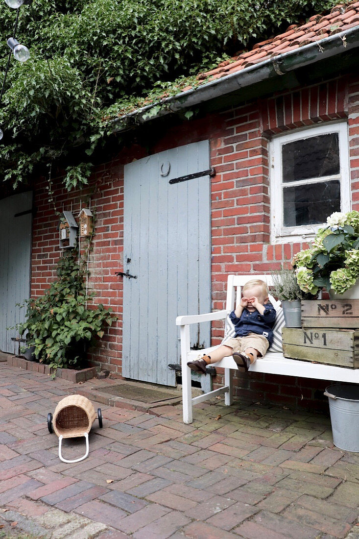 Junge auf einer Gartenbank neben Holzkisten vorm Backsteinhaus