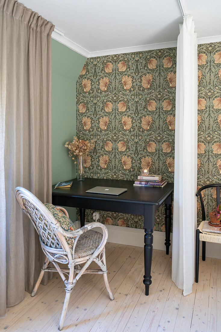 A black desk with a wicker chair behind a curtain in guest room with wallpaper