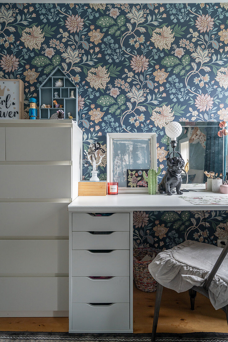 A desk and a chest of drawers in front of floral wallpaper
