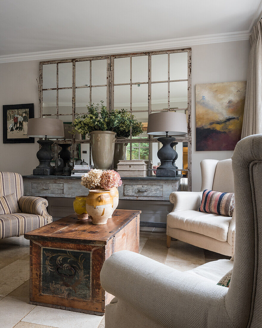 Pair of large grey lamps on large french console table in seating area with wing-back armchairs and antique chest