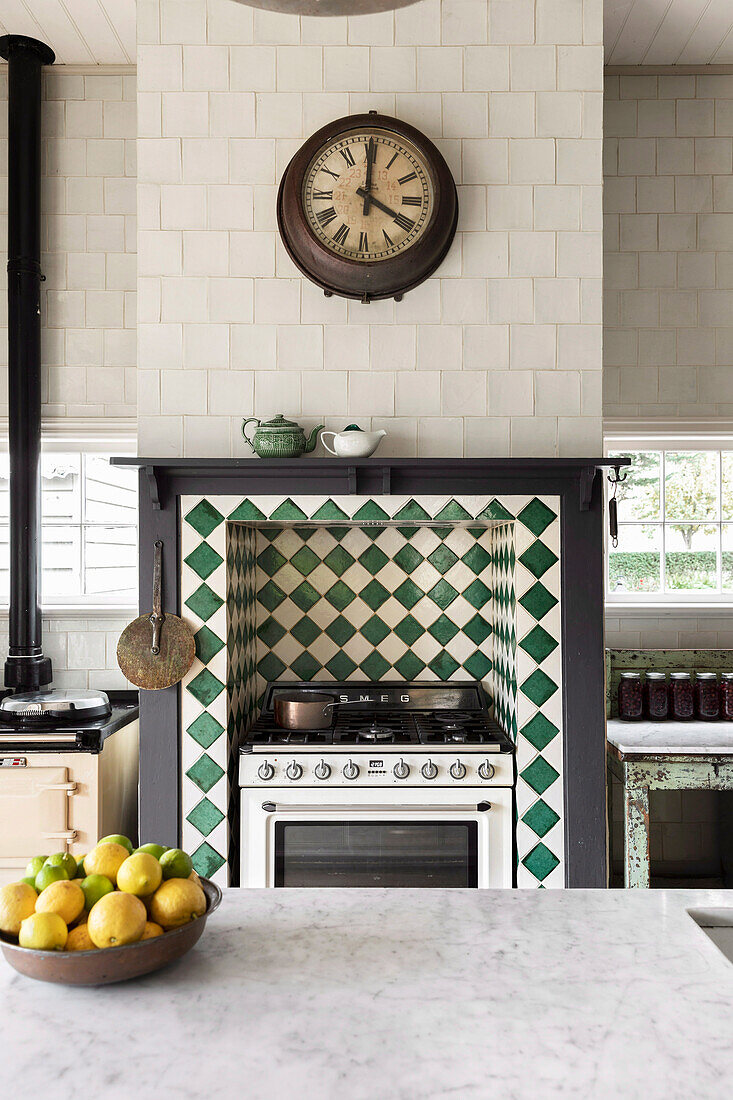 Blick über Kücheninselm mit Marmor-Arbeitsplatte auf Gasherd in Wandnische mit grün-weißen Fliesen, darüber Küchenuhr