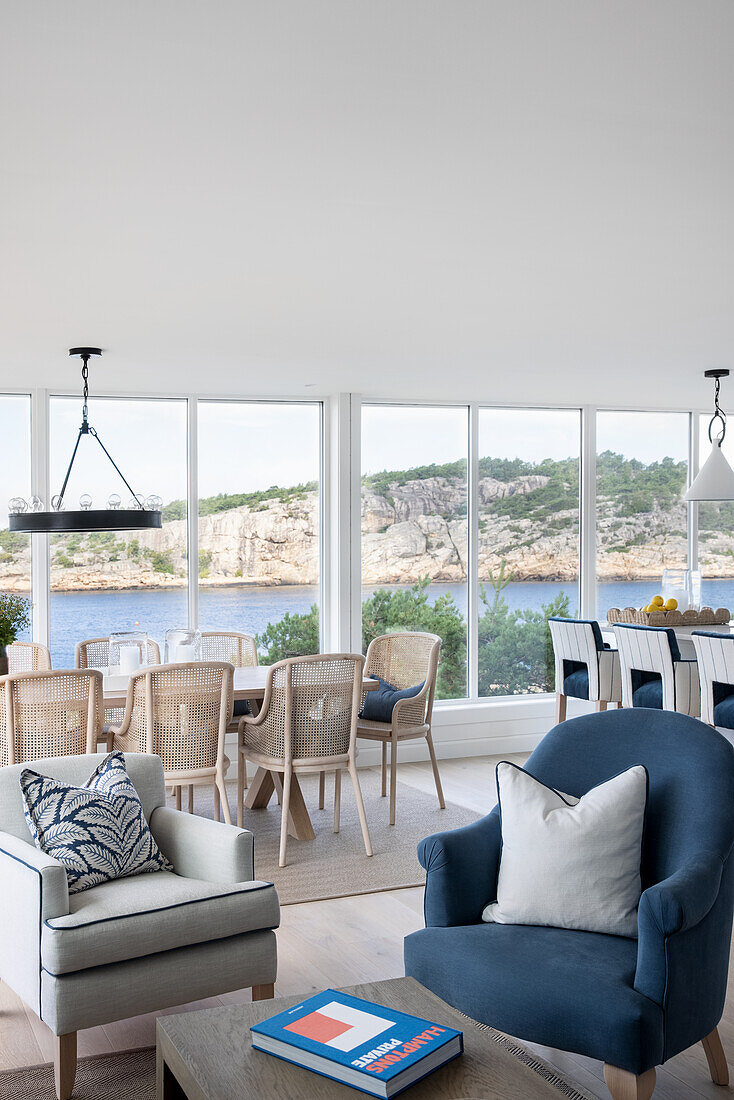 Open-plan living room with seating area and dining area in front of wall of windows, view of coastal landscape
