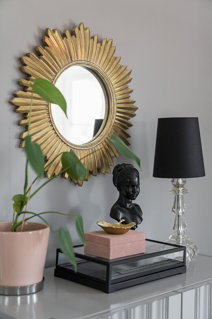Console with decorative objects, table lamp and houseplant, sun mirror above table