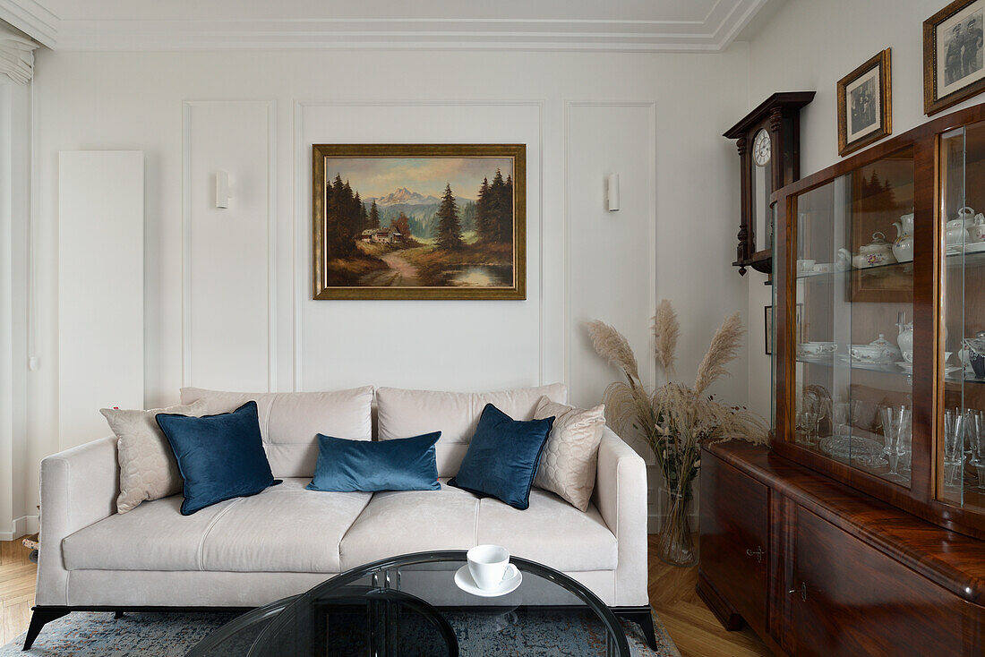 Light-colored sofa with throw pillows in front of antique sideboard, painting on white wall