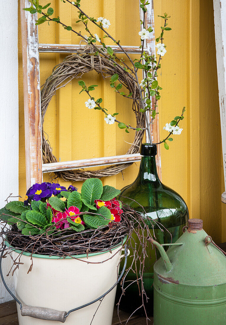 Emailleeimer mit Primeln (Primula), Ballonflasche mit Obstzweigen und Kranz aus Clematiszweigen an Hauswand