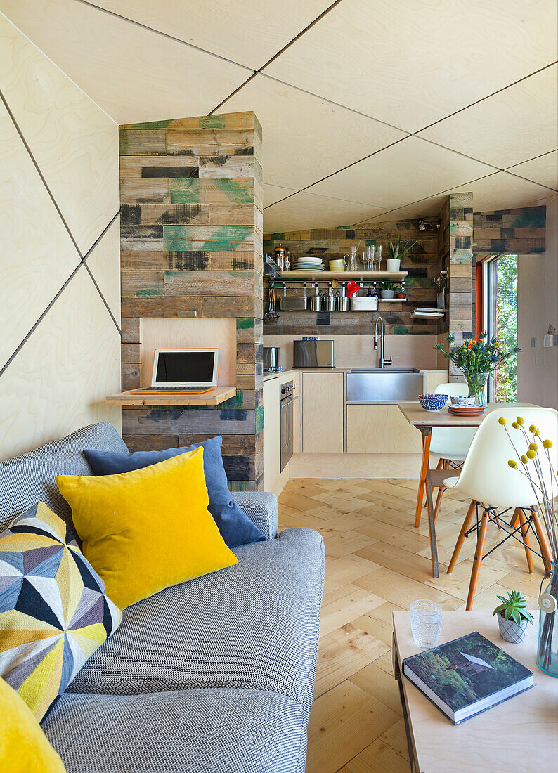 Heller, offener Wohnraum mit grauem Polstersofa, kleinem Essbereich und Küche mit Wänden aus Holzkisten, rautenförmige Decke mit LED-Leisten