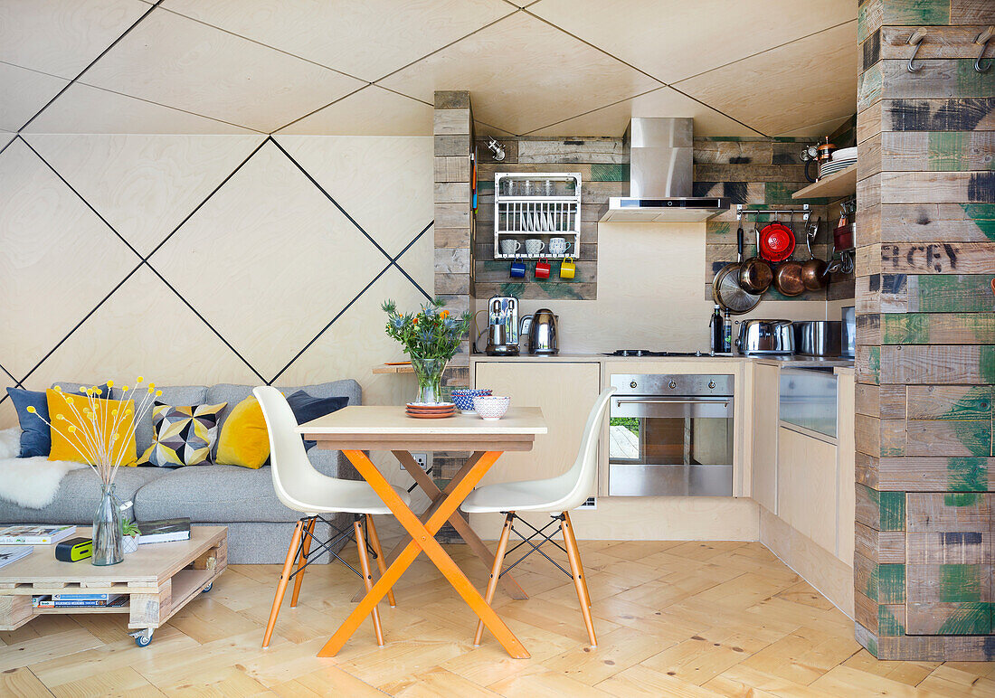 Heller, offener Wohnraum mit kleinem Essbereich und Küche mit Wänden aus Holzkisten, rautenförmige Decke mit LED-Leisten