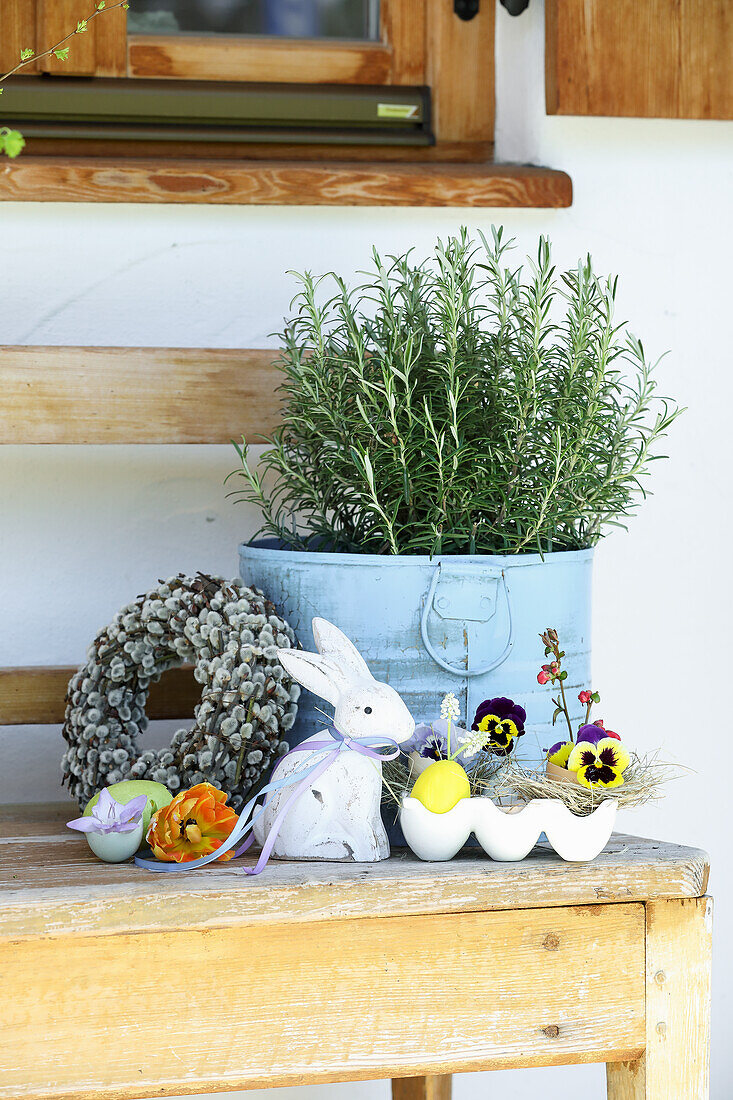 Rosemary pot surrounded by Easter decorations with pussy willow wreath, Easter bunny and pansies in an egg carton