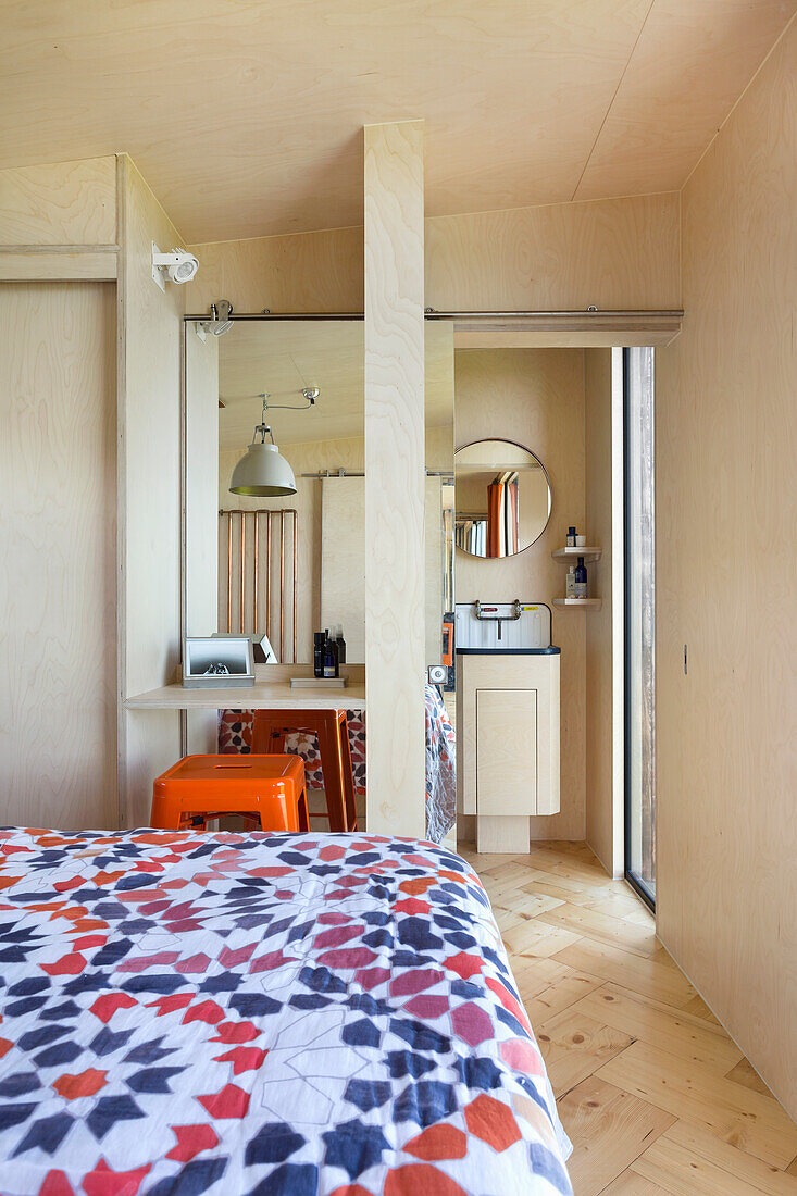 Doppelbett mit bunter Tagesdecke, kleiner Kosmetiktisch, dahinter verspiegelte Schiebetür und Blick ins Badezimmer