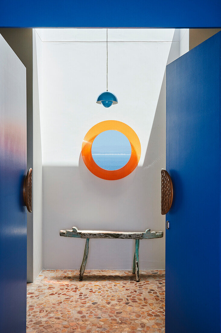 Blick durch geöffnete, blaue Flügeltüren in Einganshalle mit weißer Wand, orangefarbenem Bullauge und rustikaler Bank auf Steinboden