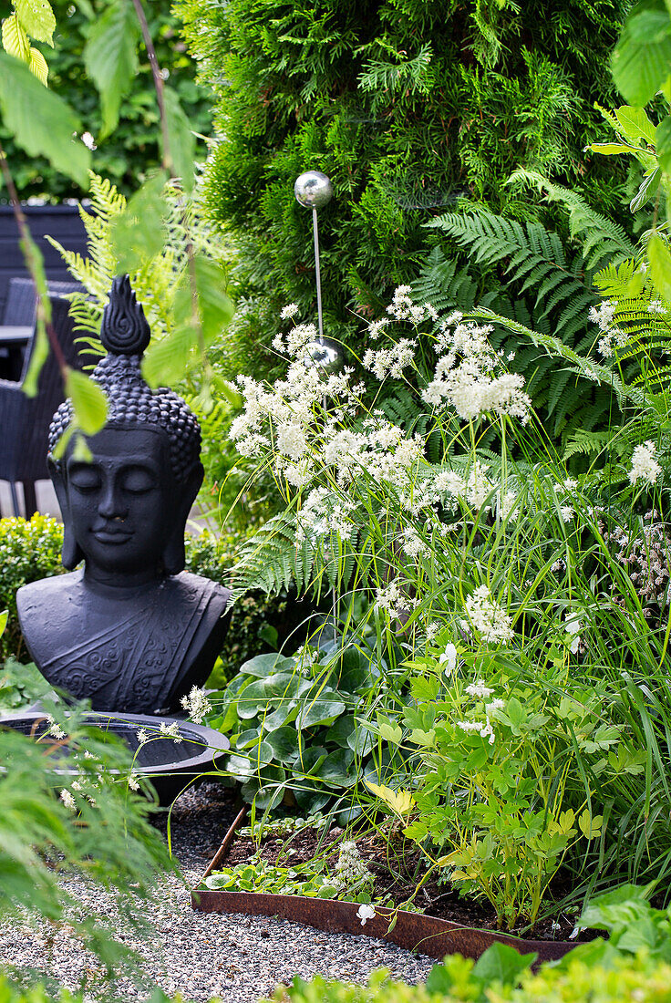 Buddha-Statue umgeben von üppiger Gartenbepflanzung und Dekorelementen