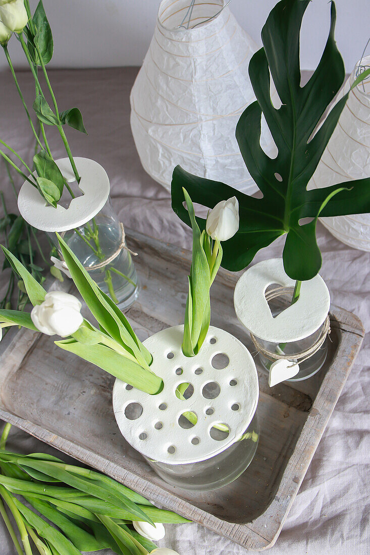 Decorative vases with white tulips and monstera leaf on wooden tray