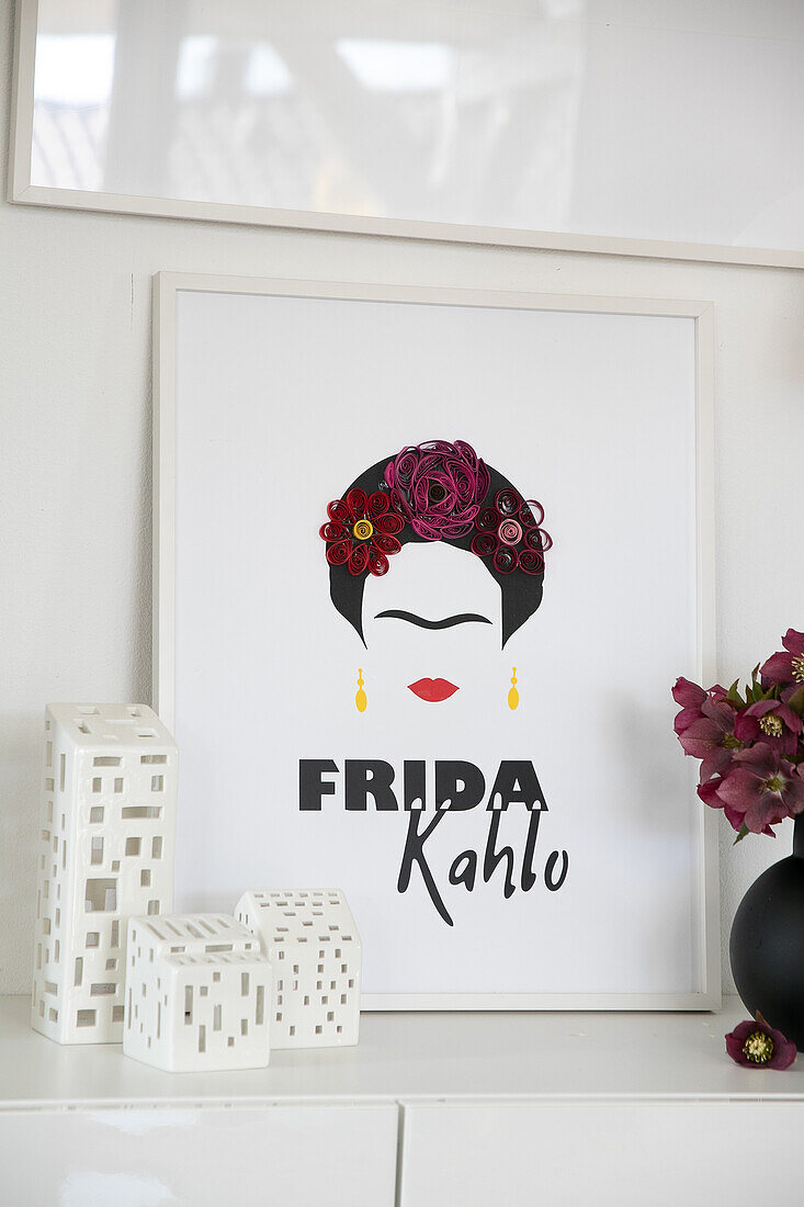 Bild mit Frida Kahlo-Motiv auf weißem Sideboard neben Blumenvase und Teelichtern