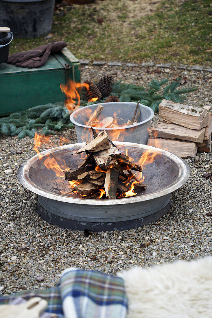 Grillfeuer in einer Feuerschale, daneben Zinkschüssel mit kleinem Brennholz und Holzscheite