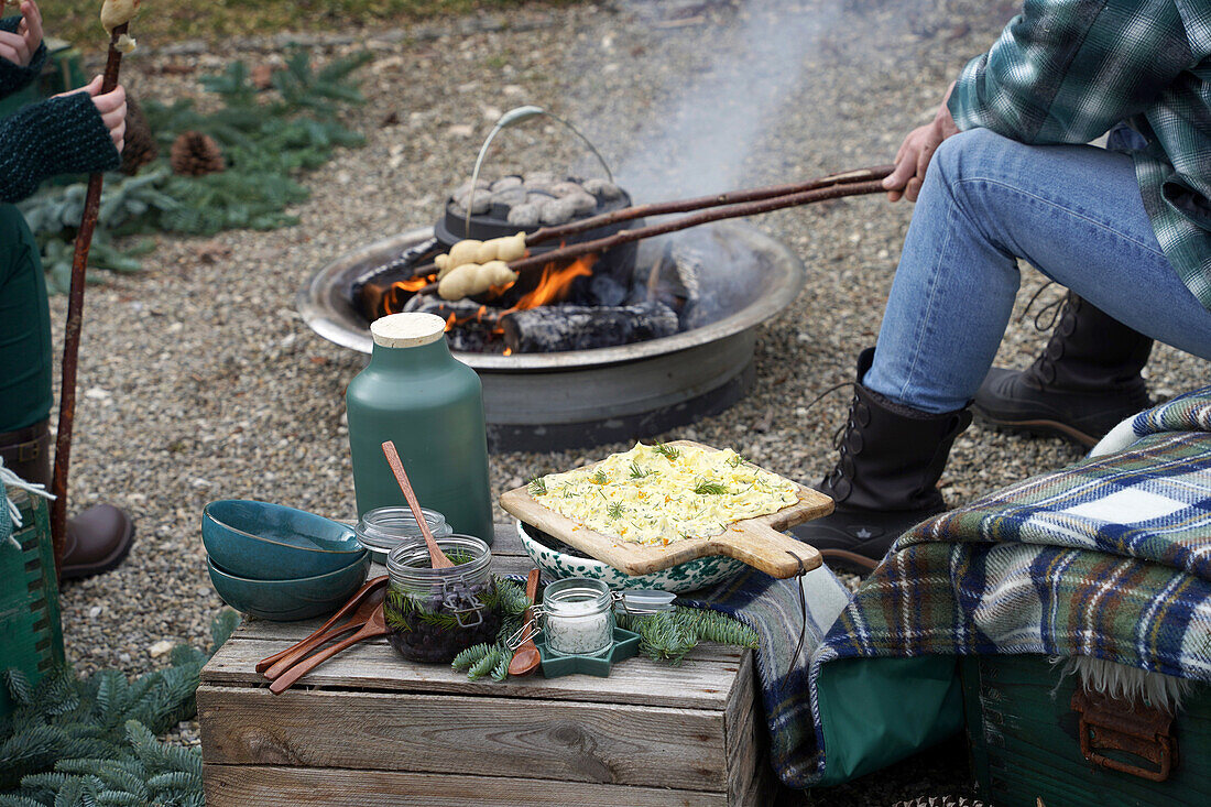 Gemütliche Outdoor-Mahlzeit am Feuer mit Stockbrot, Holzkiste mit Geschirr, Schlehenoliven, Fichtensalz und Butterbrot mit Fichtenbutter auf einer Holzkiste