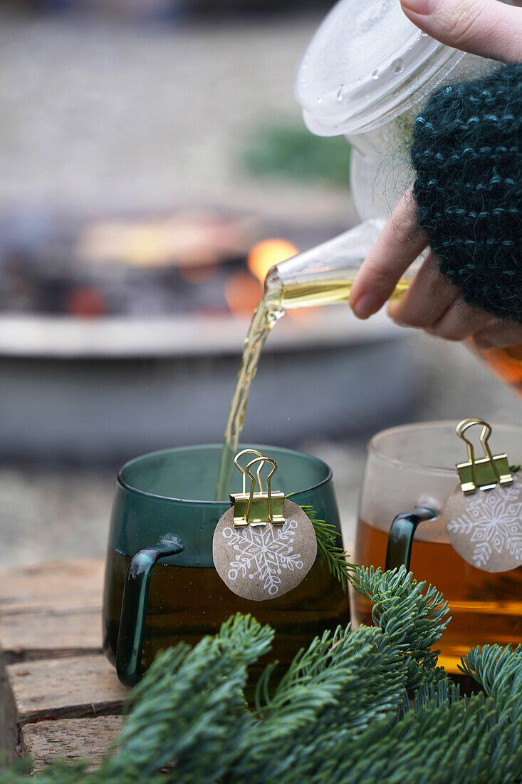 Woman pours tea in cups with tag, snowflake motif made of sustainable wrapping paper, next to fir branches, wintery terrace in front of fire pit