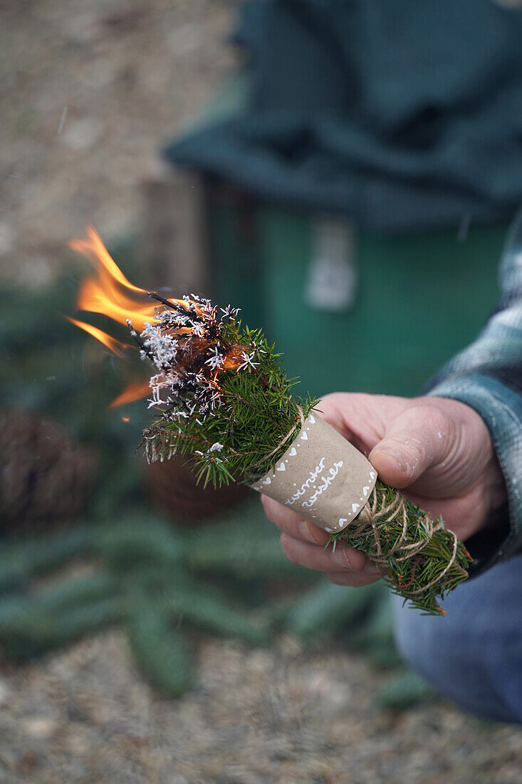 Man holding a burning bundle of incense, DIY incense bundle made of juniper and spruce branches with 'winter wishes' label