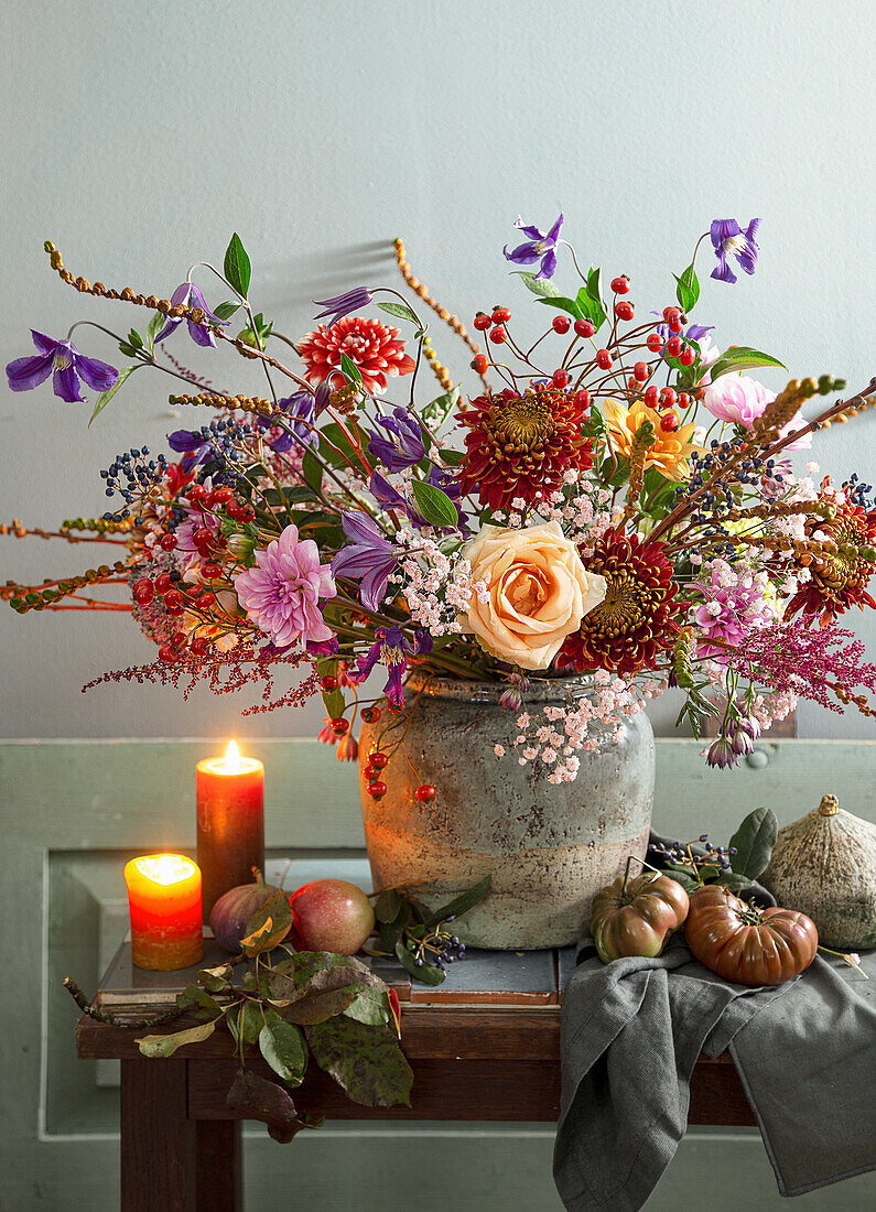 Autumn bouquet in a stone vessel surrounded by apples, tomatoes and burning candles