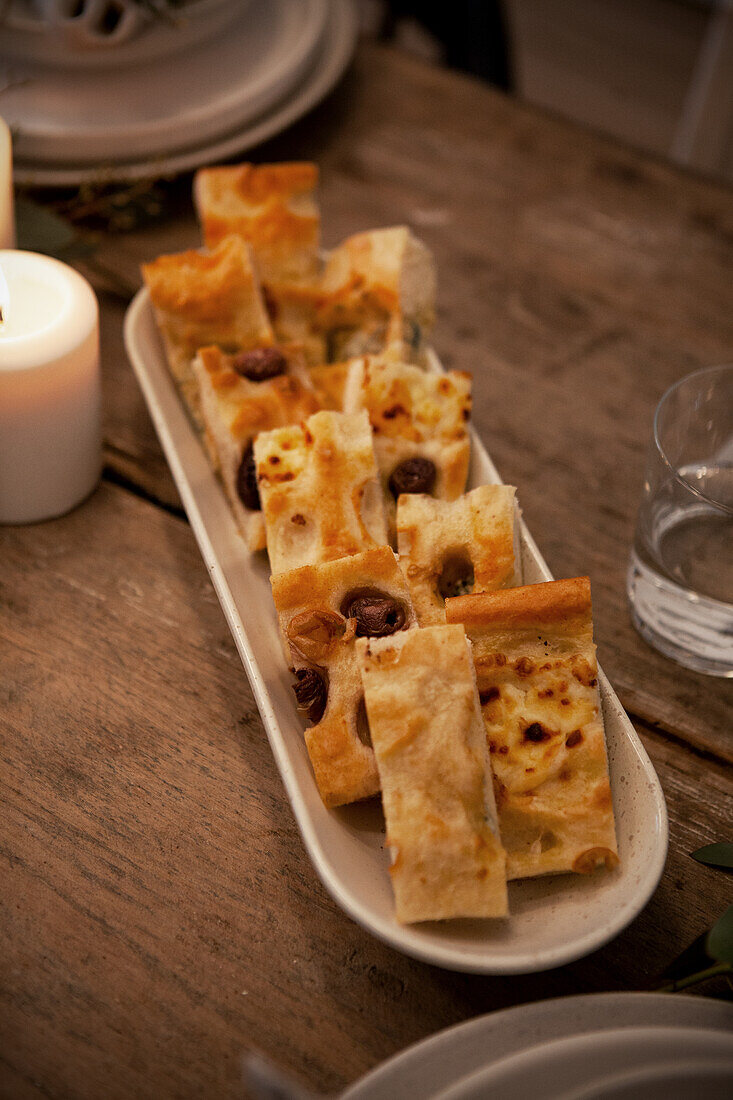 Focaccia mit Oliven auf länglicher Servierplatte neben Kerzenlicht