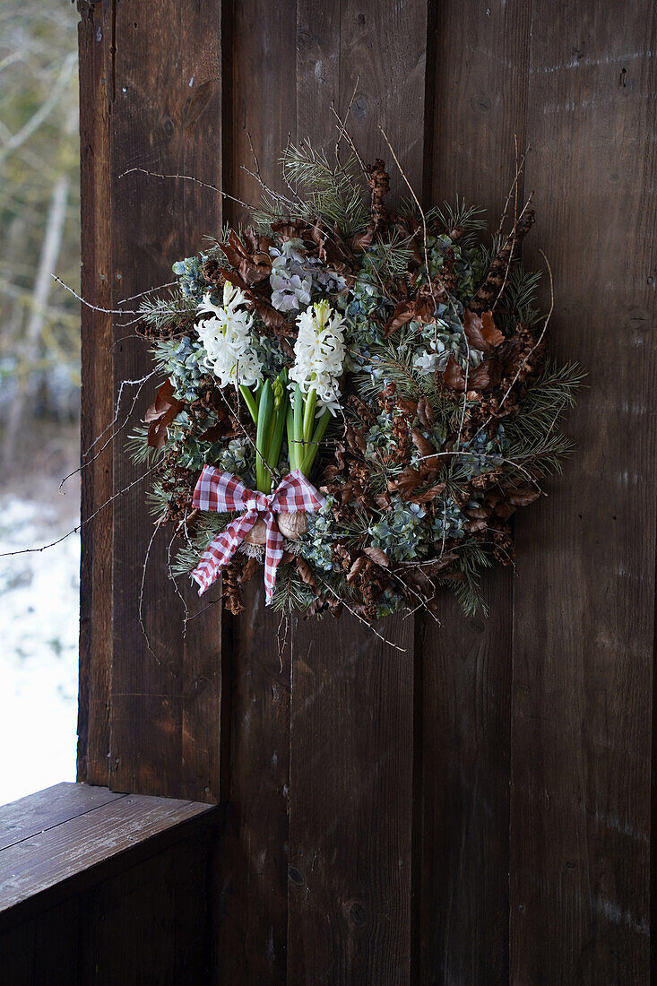 Winterkranz aus Naturmaterialien wie Lärchenzweige, Kiefernzweige, getrockneten Hortensien, Rotbuchenblätter und Farnzweige, weiße Hyazinthen, an einer Holzwand hängend