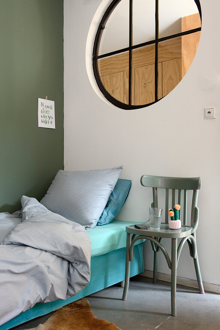 Schlafzimmer mit Einzelbett, rundem Fenster, grüner Wand und grünem Holzstuhl als Nachttisch