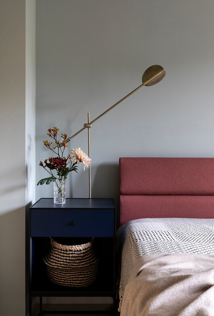 Bedside table in blue with flower arrangement and brass wall lamp next to bed