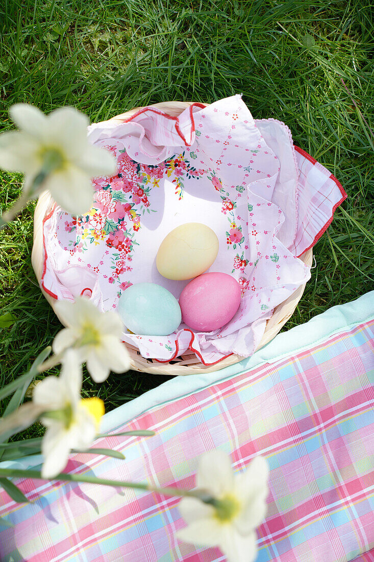 Coloured Easter eggs in a basket next to a chequered cloth in the grass