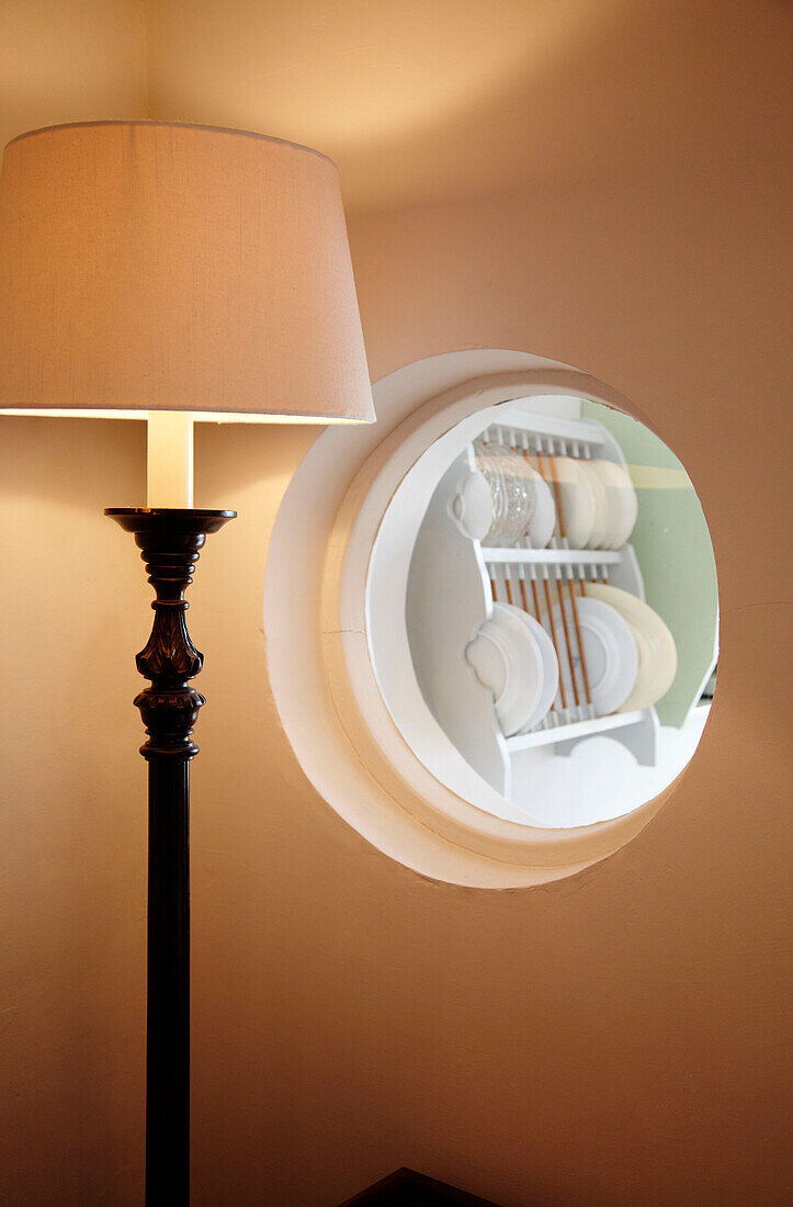 Plate rack viewed through porthole window with lit standard lamp in Brighton home East Sussex, England, UK