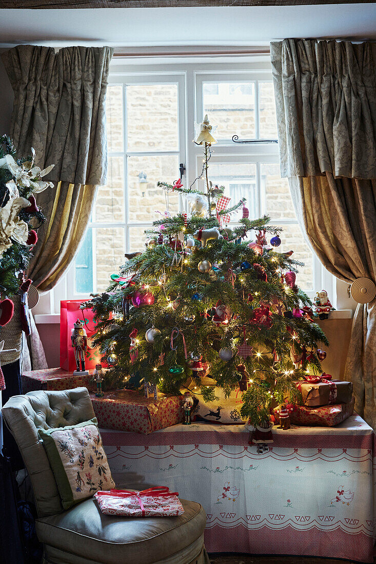 Weihnachtsbaum mit beleuchteten Lichtern und Geschenken im Fenster eines Landhauses, UK