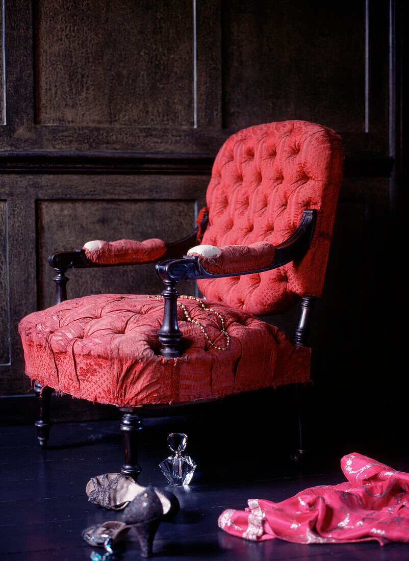 Ein abgenutzter Sessel im Vintage-Stil in einem dunkel getäfelten Raum mit Kleidung und Schuhen auf dem Boden
