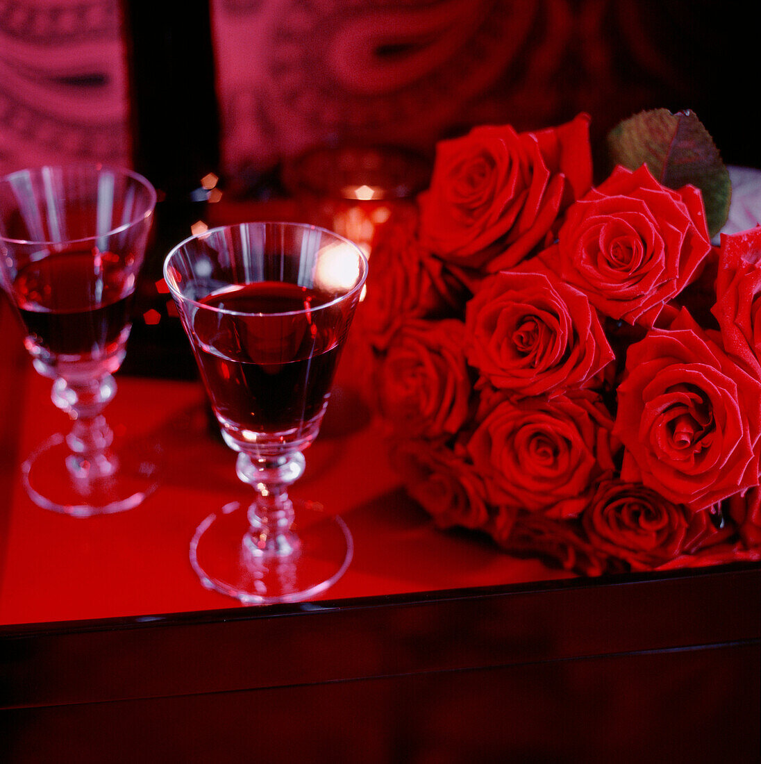 A bunch of red roses resting on a tabletop next to glasses of red wine