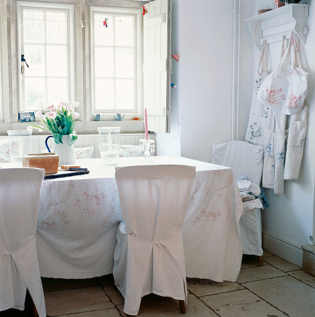 Weißes, helles Esszimmer mit losen Bezügen auf den Esszimmerstühlen und Tischtuch auf dem Tisch, Steinboden und ein großes Fenster