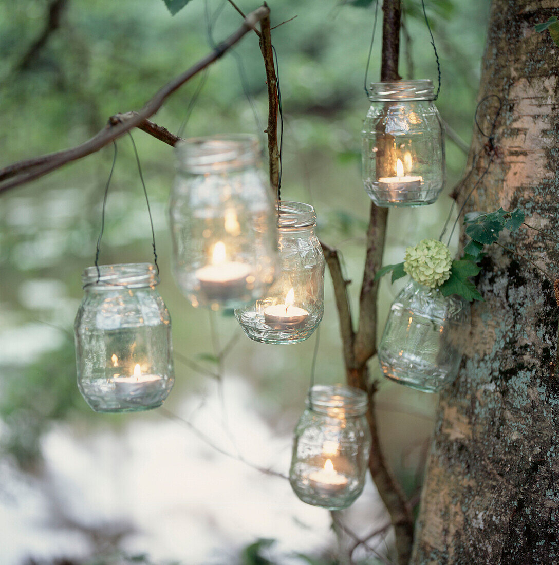 Recycled glass jam jars reused as candle holders hanging from tree branches in a wooded garden